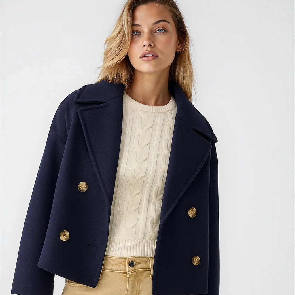 Woman wearing a navy blue coat with gold buttons, white sweater, and beige pants on a white background